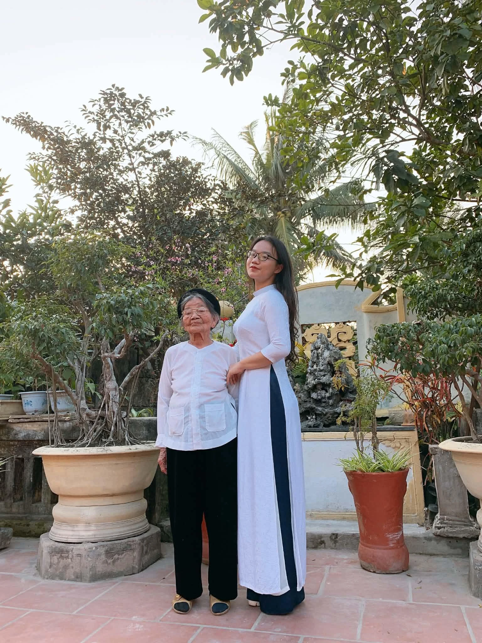 Con gái trong tà áo dài trắng đứng bên bà dưới tán cây trong sân nhà